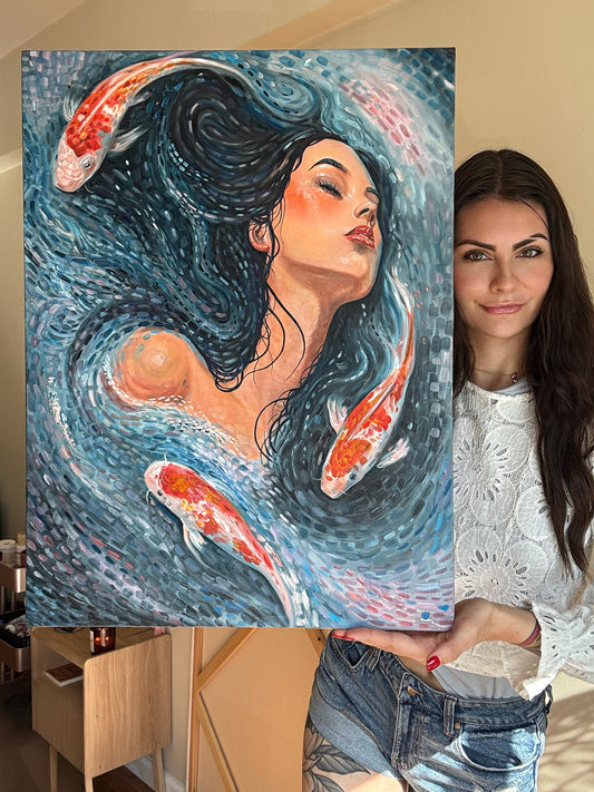 A woman holds up a large canvas painting featuring a stylized woman with flowing dark hair and koi fish swimming in swirling blue water. The woman is wearing a white lace top and denim shorts, standing indoors next to the artwork.
