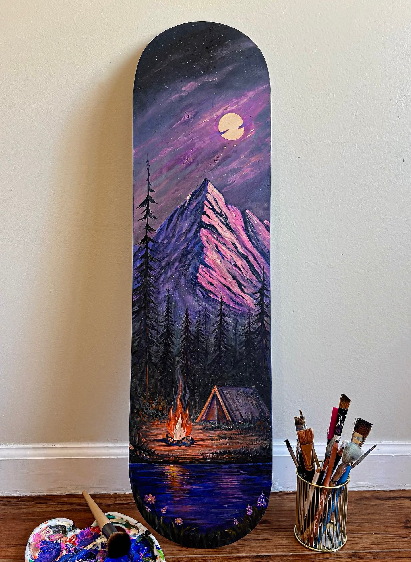 A custom hand-painted skateboard deck leaning against a beige wall on a wooden floor, featuring a scenic mountain landscape under a full moon with a campfire and tent. A paint palette and paintbrushes are visible on the floor nearby.