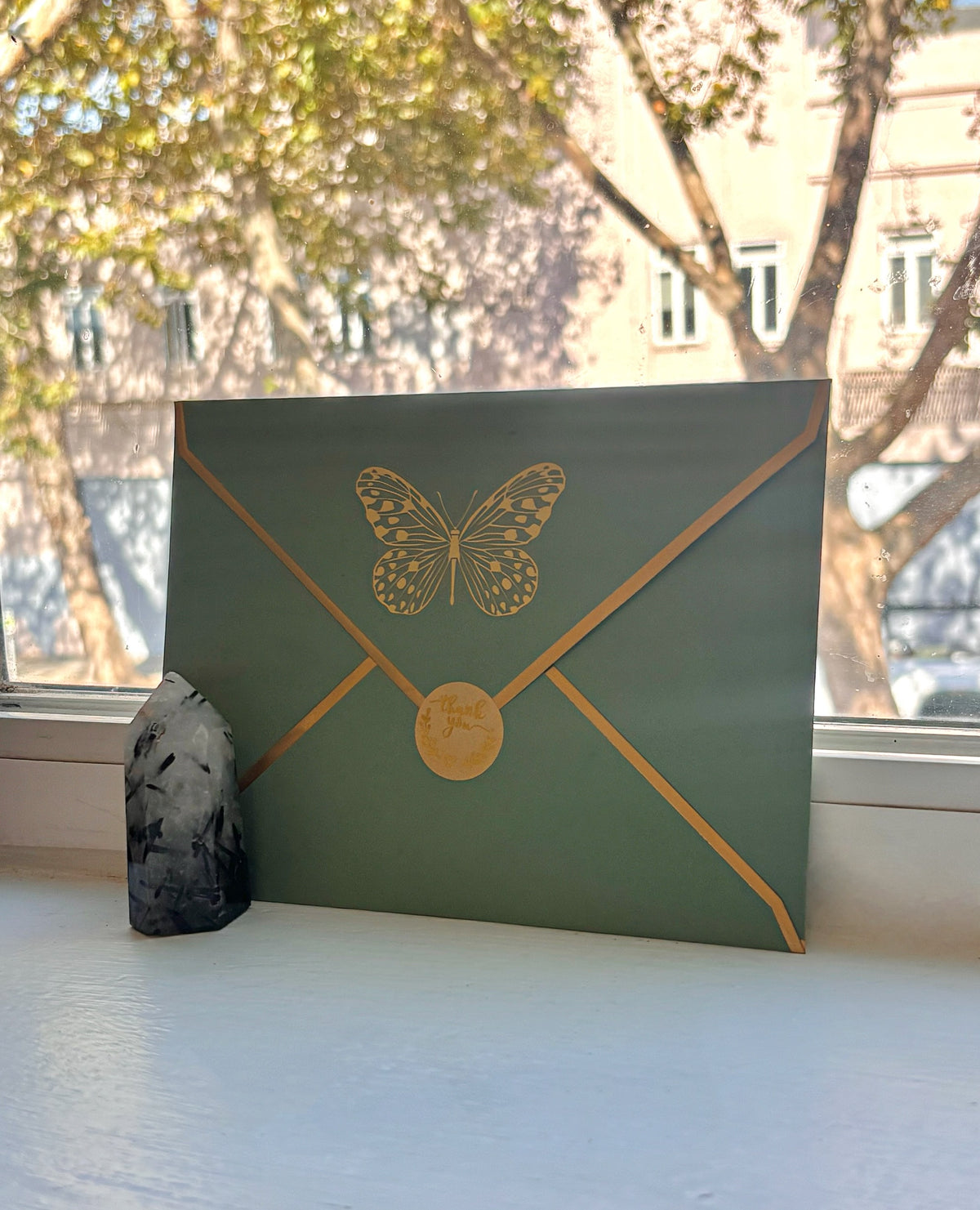 A light green envelope featuring a gold butterfly and envelope-style gold lines, leaning on a windowsill with a blurred outdoor scene behind it.