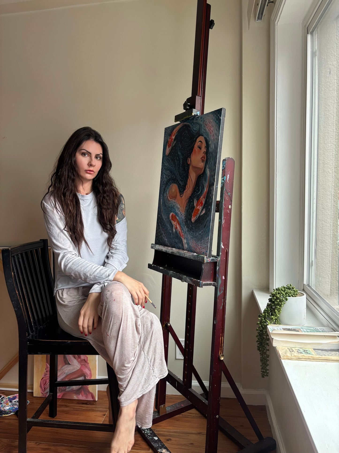 A woman with long dark hair sits on a black chair next to an easel holding a painted portrait, in a room with beige walls, a window, and a small potted plant.