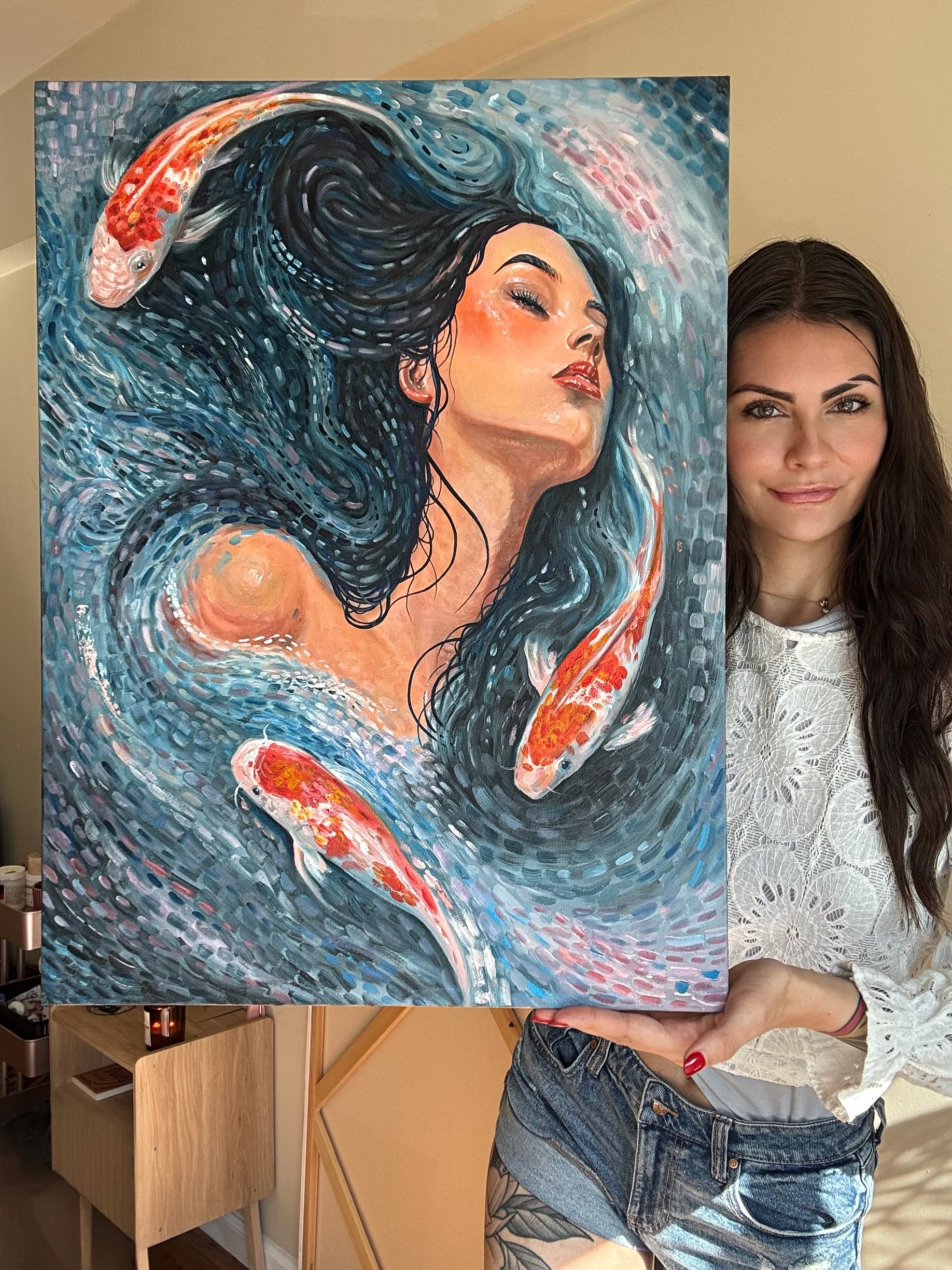 A woman holds up a large canvas painting featuring a stylized woman with flowing dark hair and koi fish swimming in swirling blue water. The woman is wearing a white lace top and denim shorts, standing indoors next to the artwork.