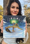 Gracey Ripa holding an original painting of a surreal, dreamlike image. A woman floating in the clouds with a tree growing from her. 