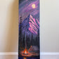 A skateboard deck featuring a painted mountain landscape at night, with a purple sky, full moon, pine trees, a campfire, and a tent near a lake, leaning against a beige wall on a wooden floor.