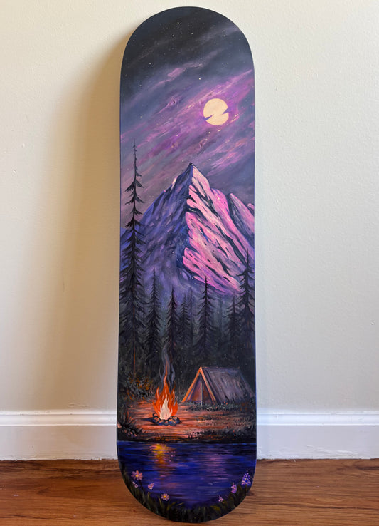 A skateboard deck featuring a painted mountain landscape at night, with a purple sky, full moon, pine trees, a campfire, and a tent near a lake, leaning against a beige wall on a wooden floor.
