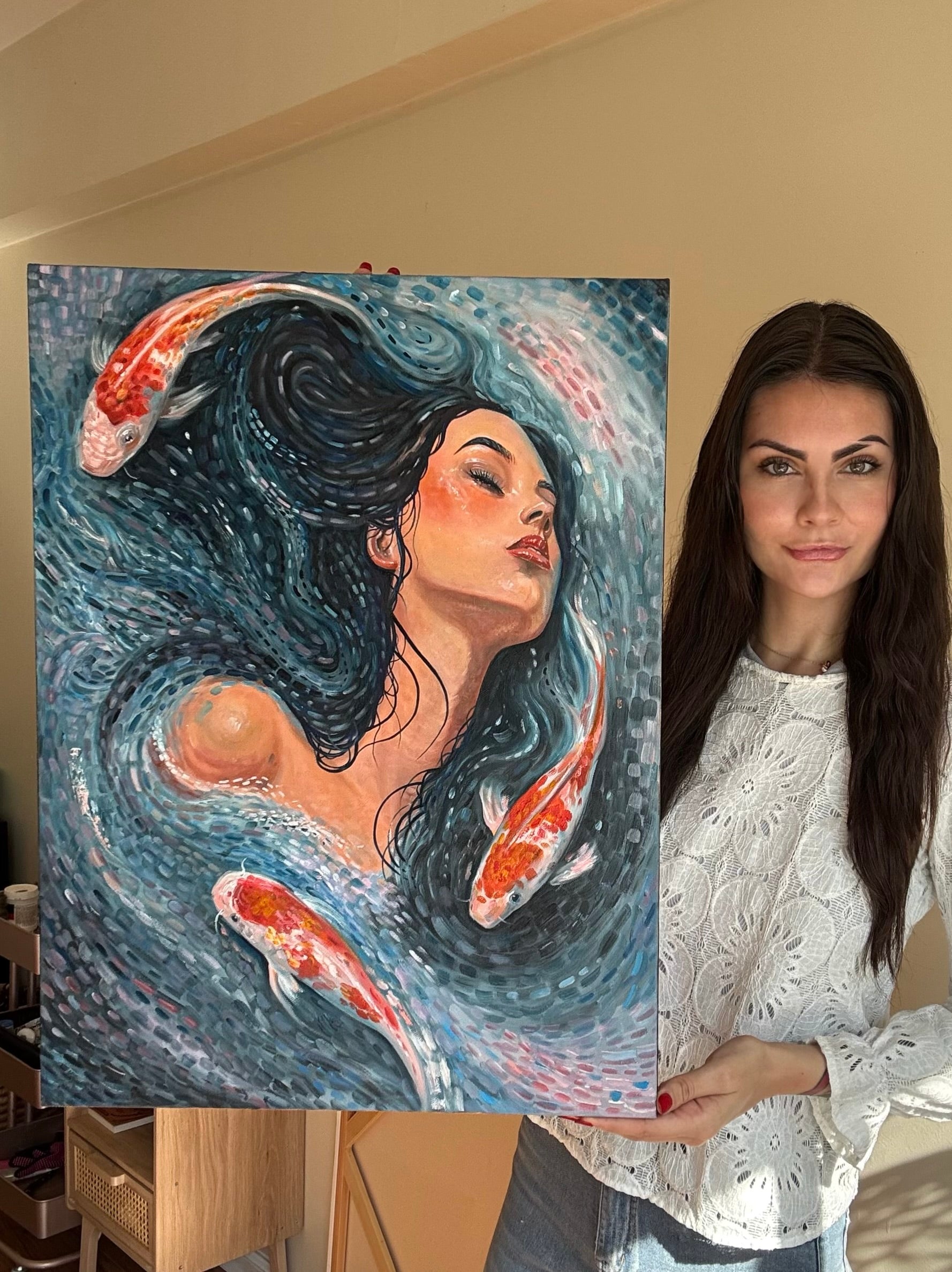 A woman stands indoors next to a large painted artwork on an easel. The painting depicts a woman with dark flowing hair surrounded by koi fish in blue water.