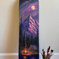 A custom hand-painted skateboard deck leaning against a beige wall on a wooden floor, featuring a scenic mountain landscape under a full moon with a campfire and tent. A paint palette and paintbrushes are visible on the floor nearby.