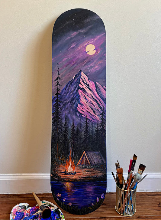 A custom hand-painted skateboard deck leaning against a beige wall on a wooden floor, featuring a scenic mountain landscape under a full moon with a campfire and tent. A paint palette and paintbrushes are visible on the floor nearby.