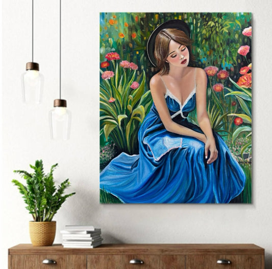 A large canvas wall art print depicting a woman in a flowing blue dress seated in a lush garden, displayed on a white wall above a wooden console table with a potted plant and books. Two hanging pendant lights are visible on the left.