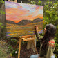 Gracey Ripa painting a landscape scene outdoors with a colorful sky and mountains.