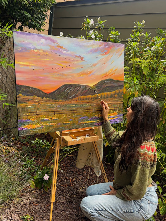 Gracey Ripa painting a landscape scene outdoors with a colorful sky and mountains.