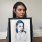 Gracey Ripa holding a framed drawing of a woman with blue hair against a plain wall.