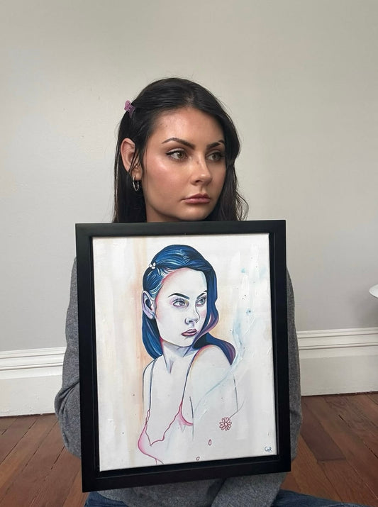 Gracey Ripa holding a framed drawing of a woman with blue hair against a plain wall.
