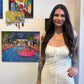 A woman with long dark hair and tattoos stands indoors wearing a white textured dress, with framed artwork and a colorful painting of a poolside scene hanging on the wall behind her.