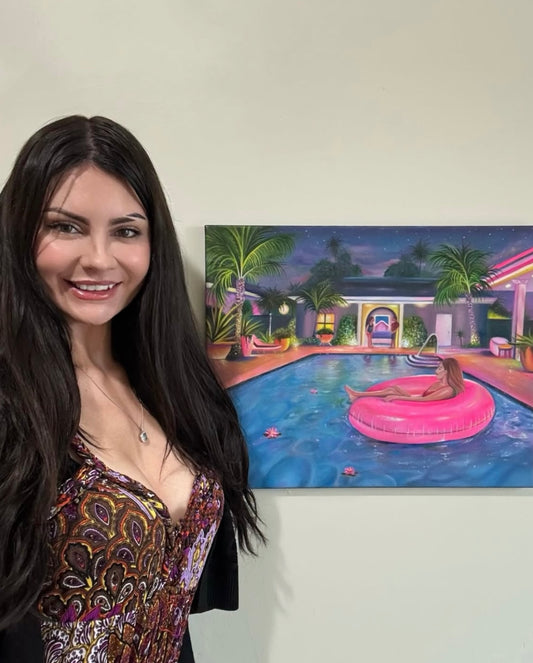 Gracey Ripa stands next to a colorful painting of a luxurious poolside scene at night, featuring a pink float and palm trees.