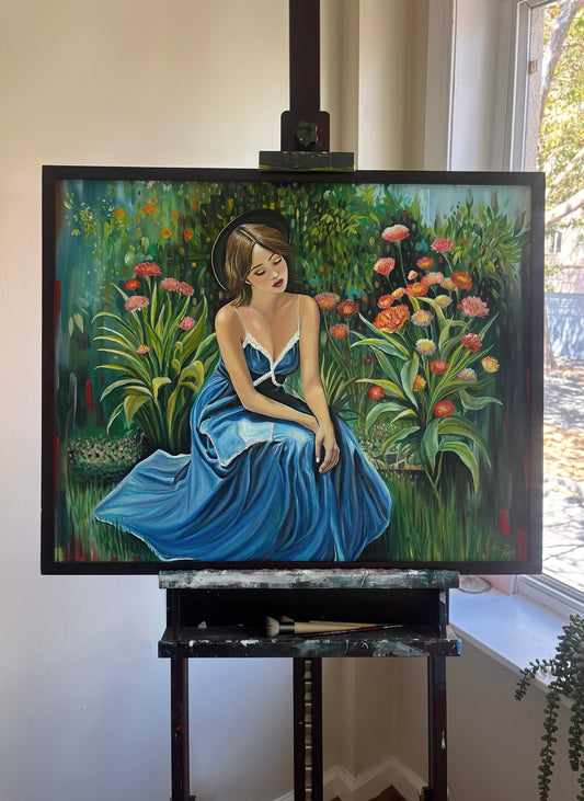 A framed painting on an easel depicting a woman in a blue dress seated in a lush garden with pink flowers. The easel stands in an indoor setting near a window.