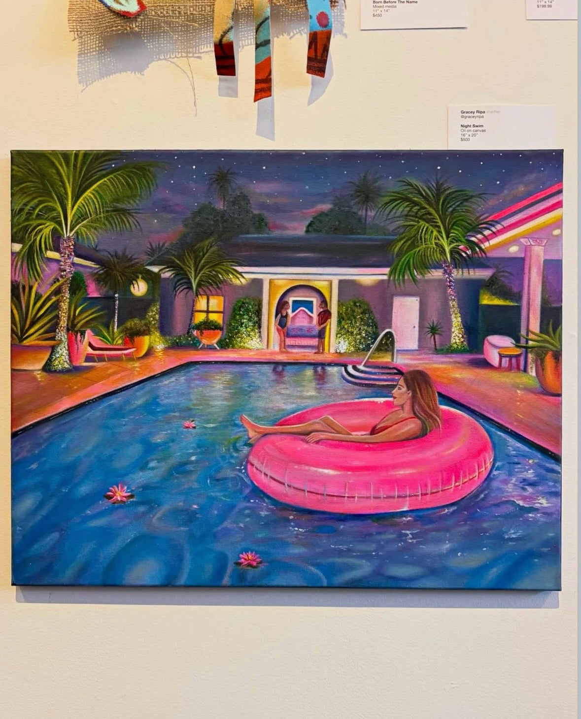 A colorful painting on canvas depicting a woman lounging in a pink float on a swimming pool at night, surrounded by palm trees and a modern house, displayed on a beige wall.