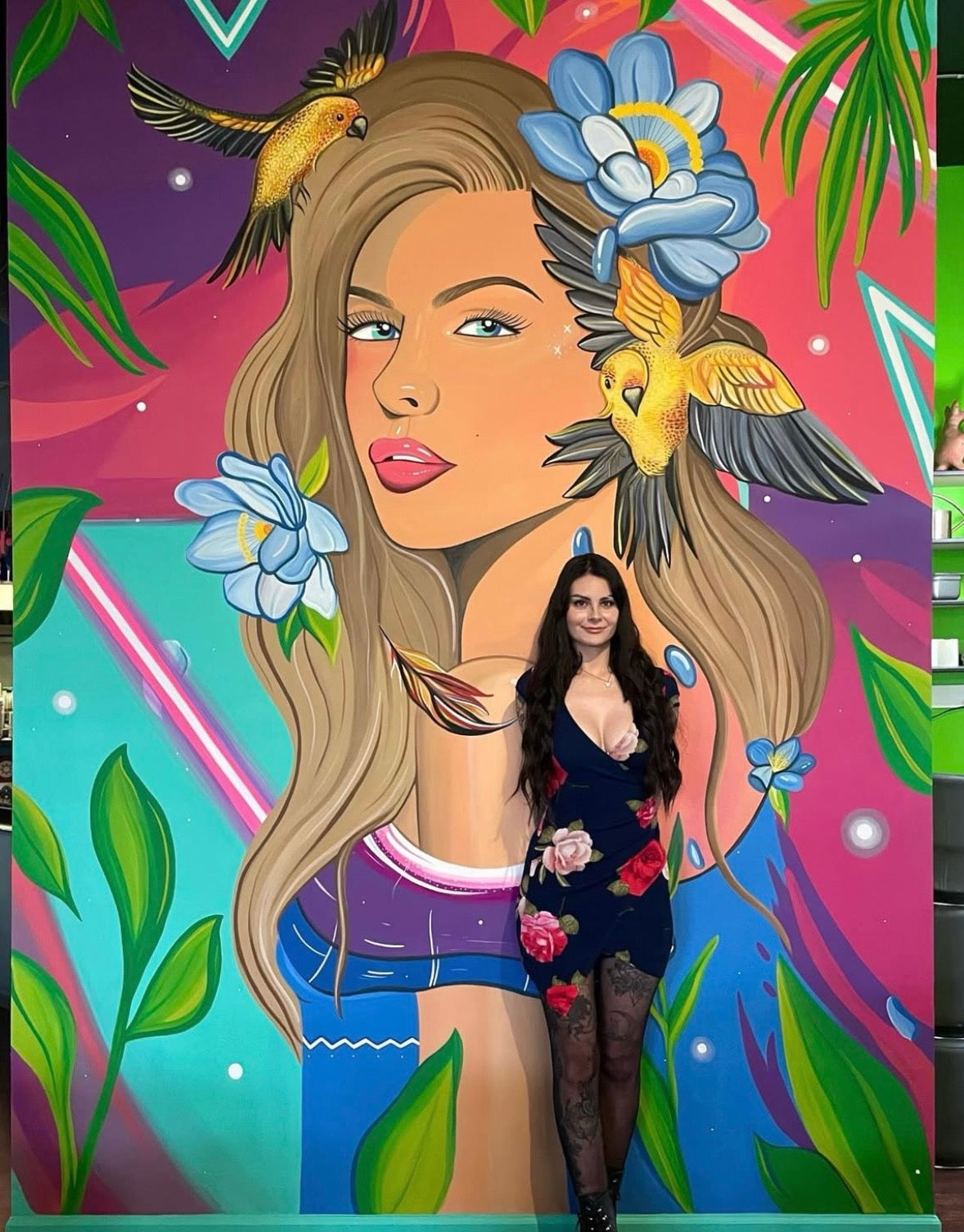 Gracey Ripa standing in front of a colorful and tropical mural of a woman with birds and flowers in her hair.