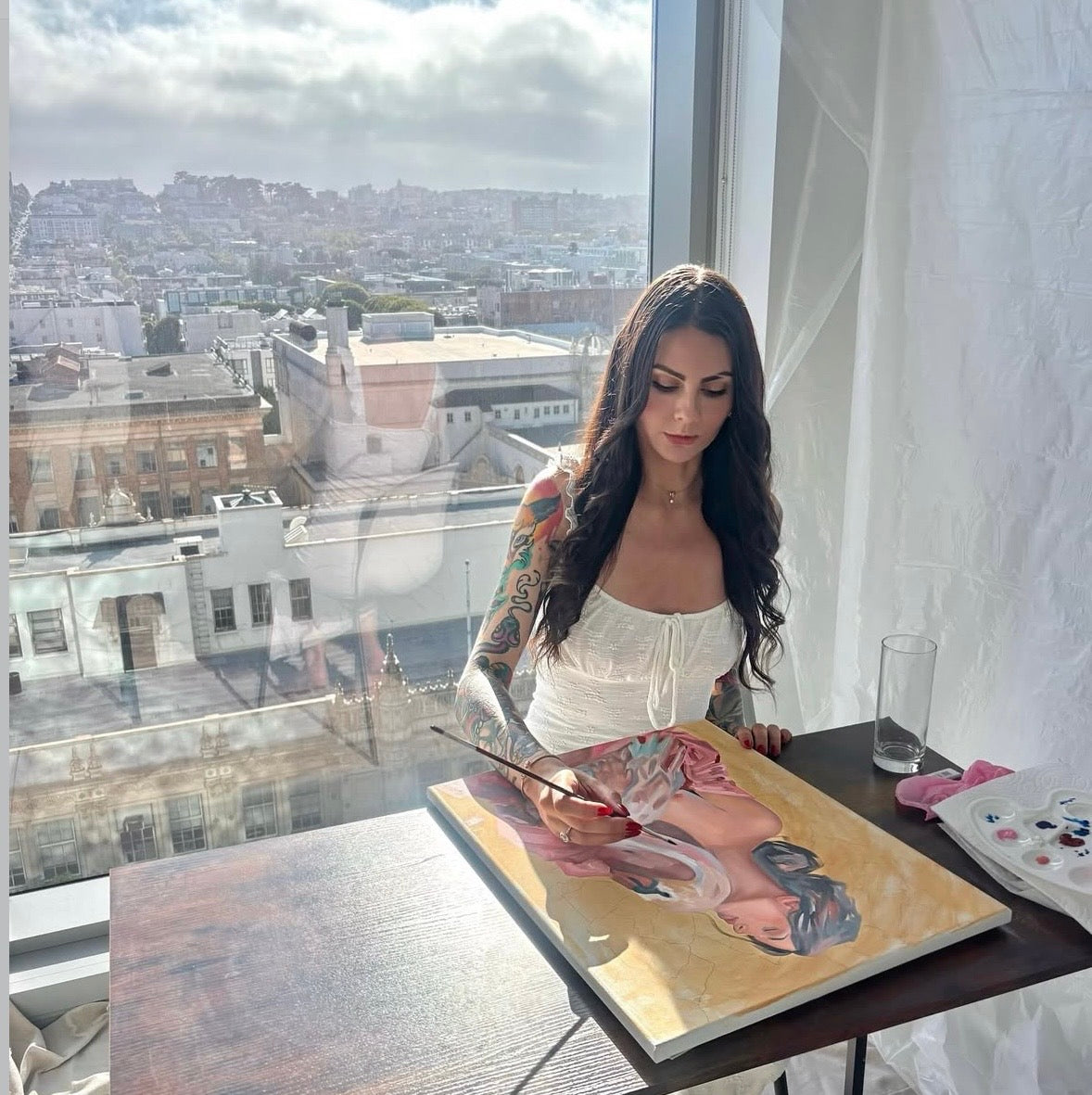 Gracey Ripa sitting and painting in an apartment with the view of San Francisco in the background. 