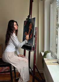 Gracey sits on a stool at an easel, painting a portrait on canvas in a bright room with a window and a small potted plant nearby.